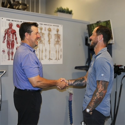 Two people shaking hands in a treatment room with anatomy charts on the wall and medical equipment nearby.