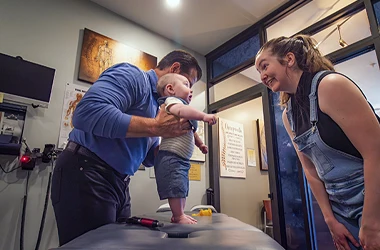Dr. Richard Kuhns is holding a baby standing on a medical table while another adult leans forward inside a clinic.